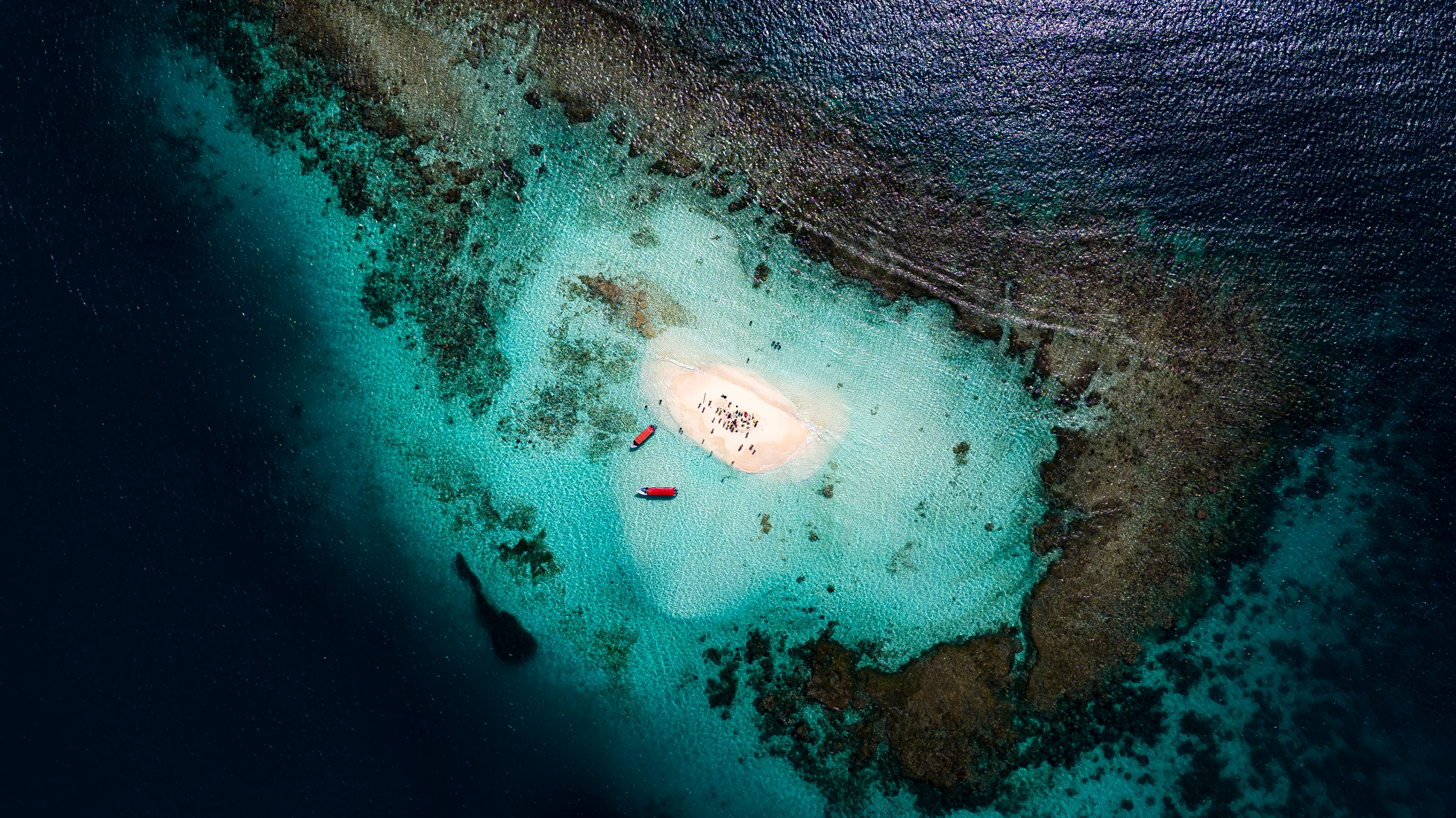 Overhead view of visitors on a white-sand sandbank with two red boats amid turquoise reef water in the San Blas Islands.