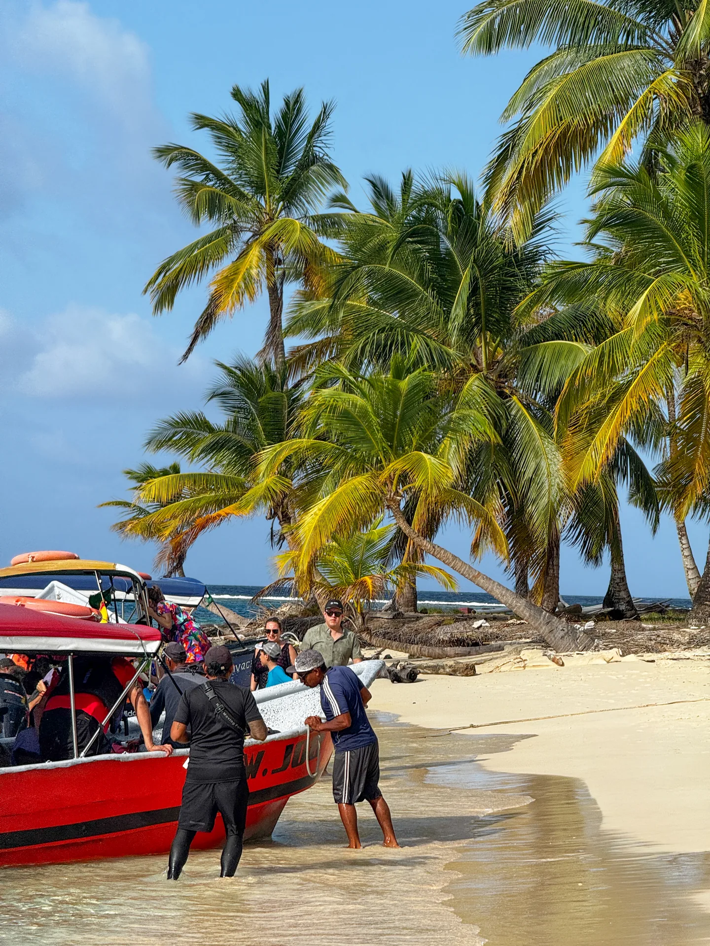 Passengers beside a red covered boat at a white-sand, palm-lined beach in the San Blas Islands.
