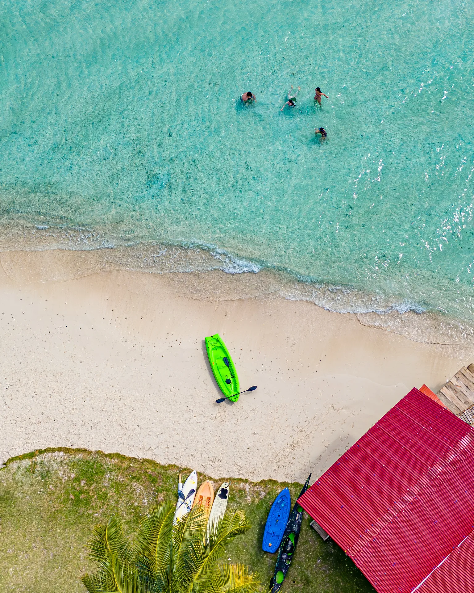 Overhead view of swimmers in turquoise water and colorful kayaks beside a sandy beach in the San Blas Islands.