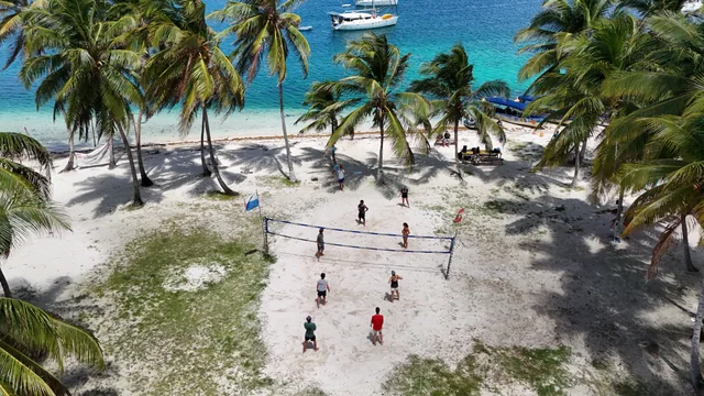 Aerial view of guests playing beach volleyball among coconut palms, with boats anchored in turquoise water in San Blas, Panama.