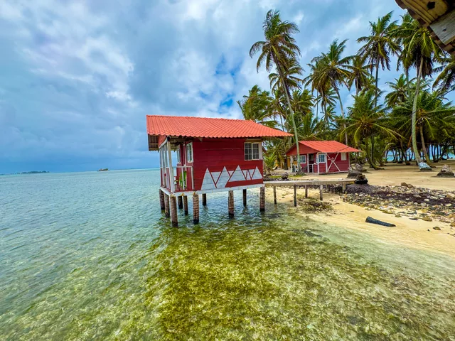 A red-and-white private cabin stands on wooden stilts above clear shallows beside Yanis Island’s palm-lined beach.